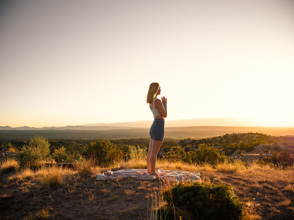 Woman in prayer pose on a hilltop blanket at sunset overlooking a vast desert landscape.